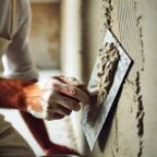 DALL·E 2024-08-27 18.07.08 - A close-up image of a person using a trowel to apply plaster or cement to a wall. The person's arm is visible, wearing a white shirt and a paint-stain